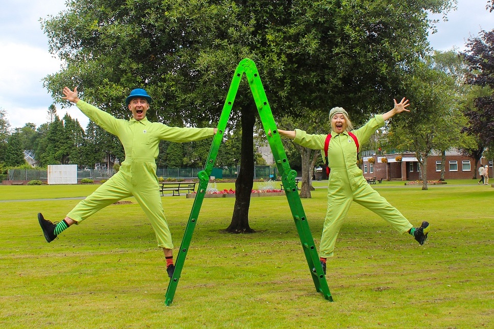 Two individuals dressed in green boiler suits, each holding onto an A-frame ladder, beam with radiant smiles on their faces.