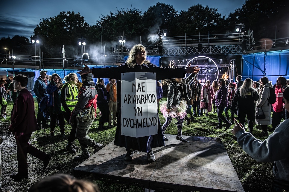 Photograph of people at an outdoor event. There is a woman standing in the middle with a sign which has the words 'Arianrhod is returning' written in Welsh / Ffotograff o bobl mewn digwyddiad tu-allan. Mae dynes yn sefyll yn y canol gydag arwydd sydd â’r geiriau Cymraeg, ‘Mae Arianrhod yn dychwelyd’