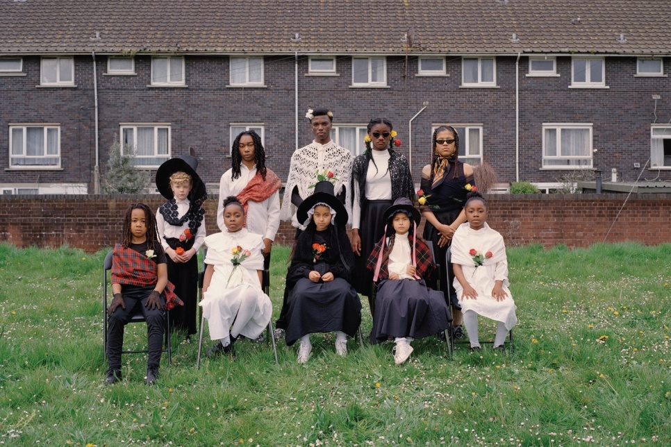 Photograph - A gathering of young people in traditional Welsh costumes standing together on grass against backdrop of a brick building / Ffotograff - Casgliad o bobl ifanc mewn gwisgoedd traddodiadol Cymreig yn sefyll gyda'i gilydd ar laswellt o flaen adeilad brics 