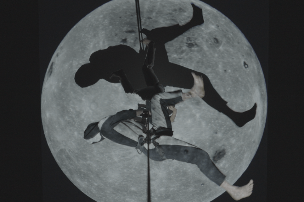 Image of two people attached horizontally to a strap in front of the moon and the night sky / Delwedd o ddau berson wedi eu cysylltu yn llorweddol â strap o flaen lleuad ac awyr y nos